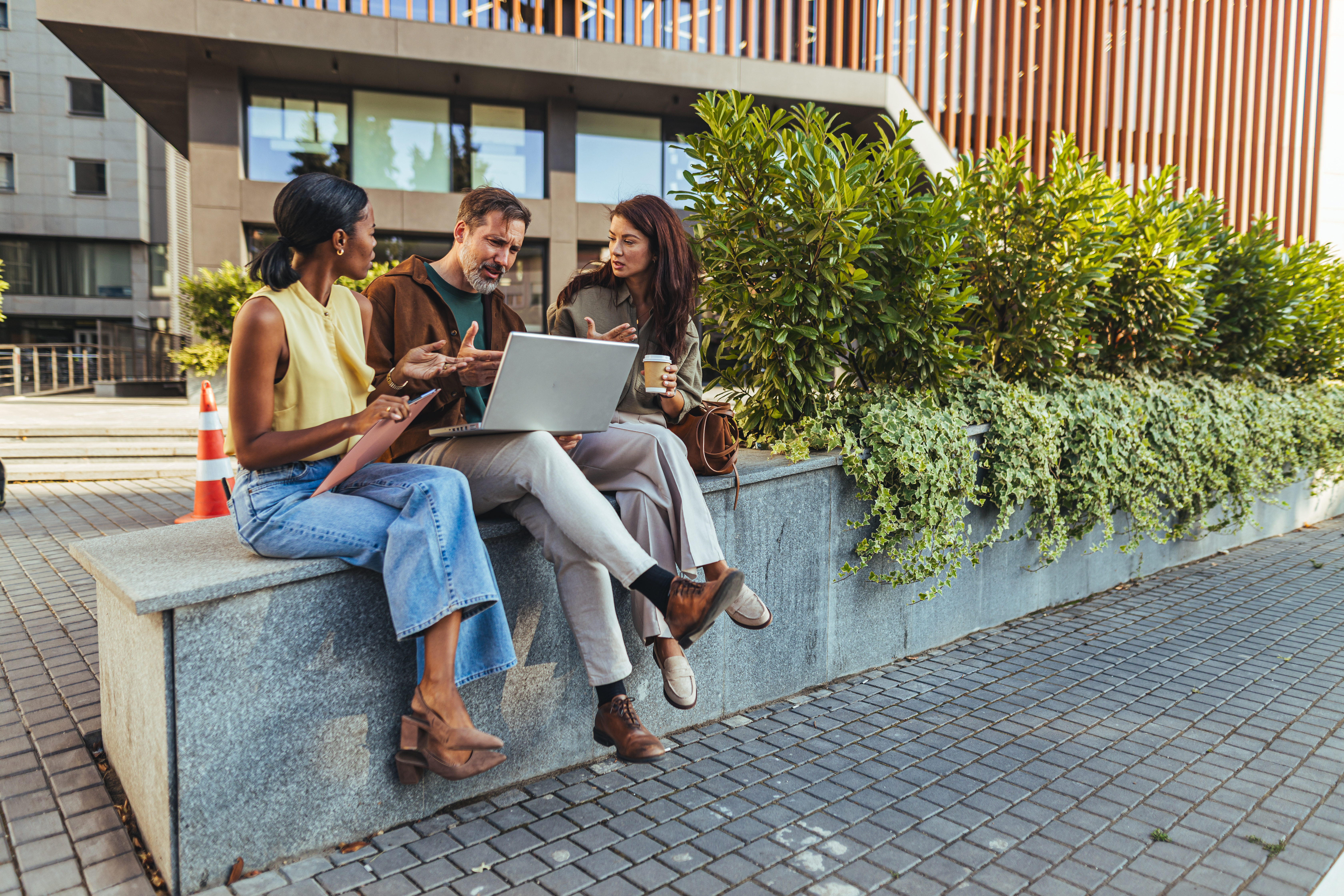 Three colleagues sit outdoors near an office building, collaborating over a laptop and coffee while discussing a project; relaxed teamwork, brainstorming and professional conversation in an urban setting.